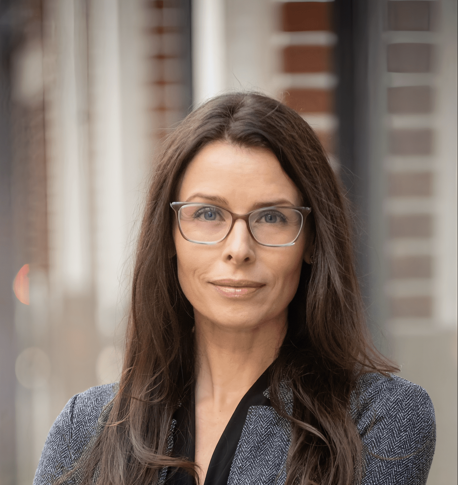 Headshot of Rebecca Phul standing in front of a brick wall with glasses and a blazer