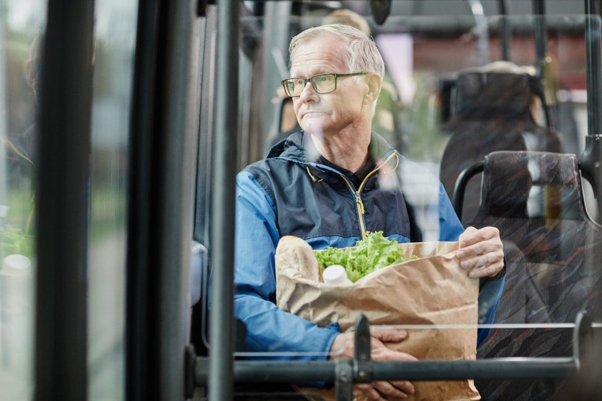 Senior man looking out window on public bus while holding bag of groceries